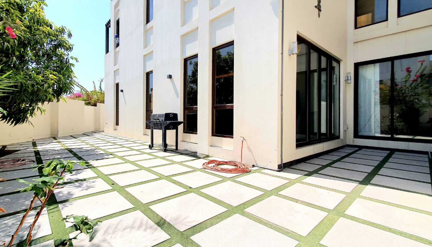 A modern house exterior with large windows, a geometric tiled patio, a barbecue grill, and a coiled hose against a white wall under a clear sky.