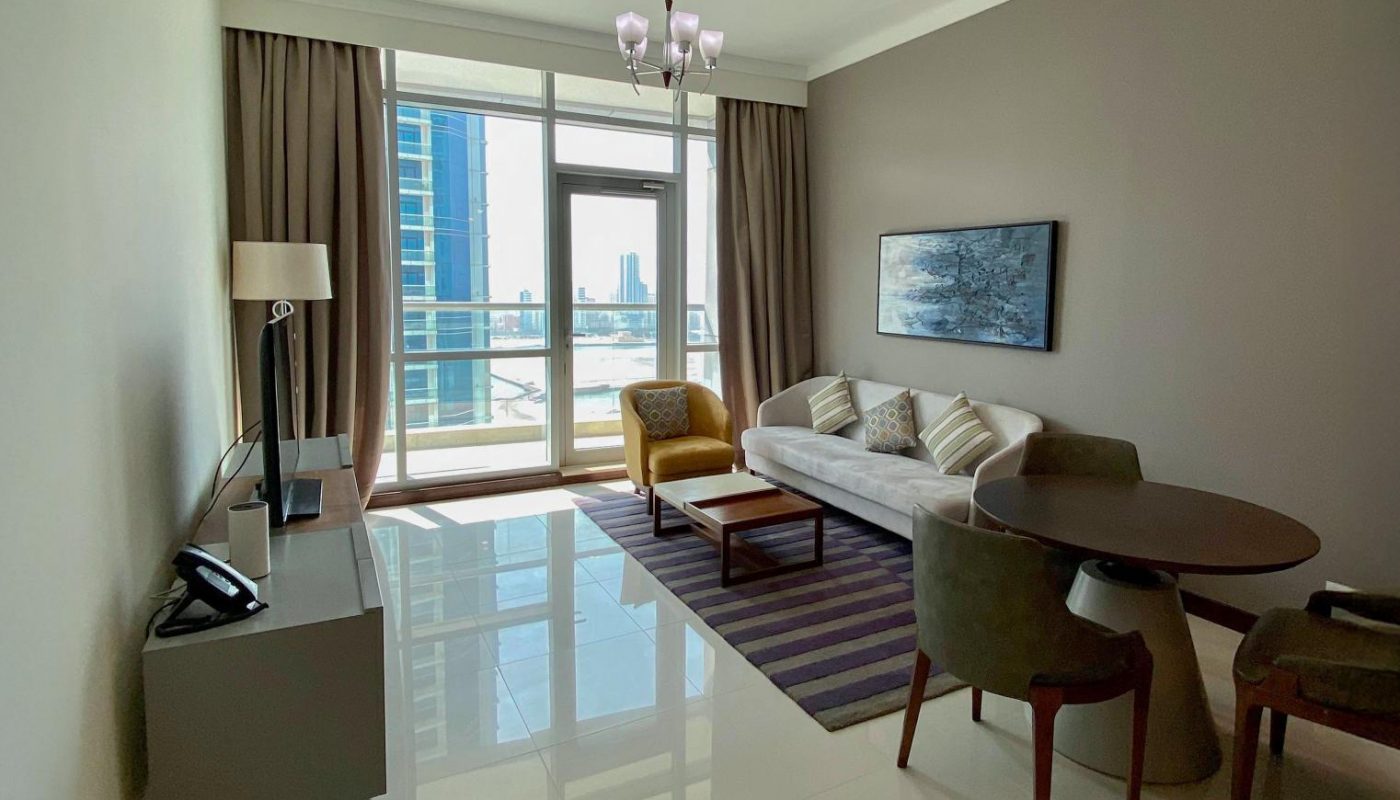 A modern living room with a sofa, coffee table, armchairs, round dining table, window with city view, and neutral decor.