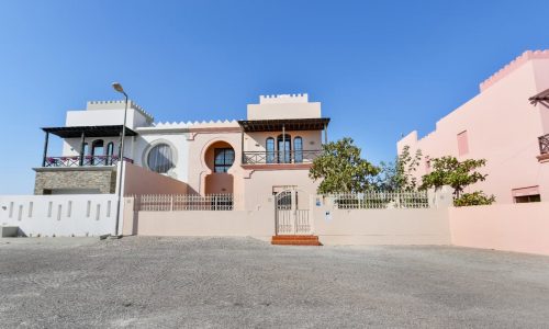 A pink and white building with a castle-like design stands against a clear blue sky, featuring arched windows, decorative balconies, and a gated entrance. The 3BR Villa in Al Areen is complemented by its expansive big garden, adding to the fairytale charm.