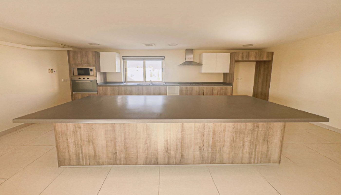 Modern kitchen with a large central island, built-in appliances, wood cabinets, white upper cabinets, and light-colored tile floor. A window is centered behind the island.