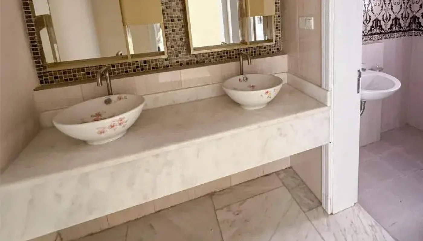 A bathroom with a marble countertop, two vessel sinks with floral designs, a mosaic-tiled mirror, and an adjacent room with a single sink.