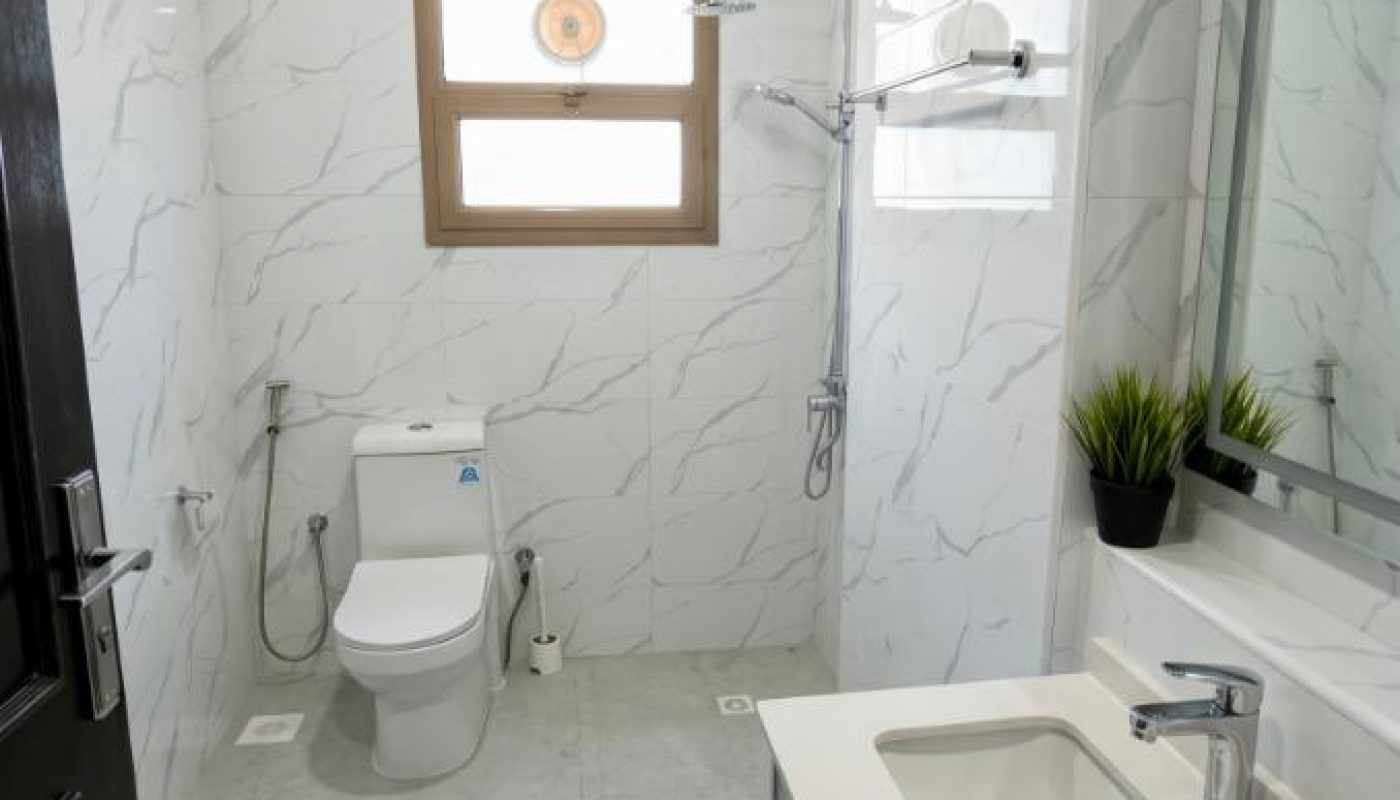 Modern bathroom with white marble tile walls, a toilet, a shower area, a rectangular sink with a mirror, and potted plants on the countertop.