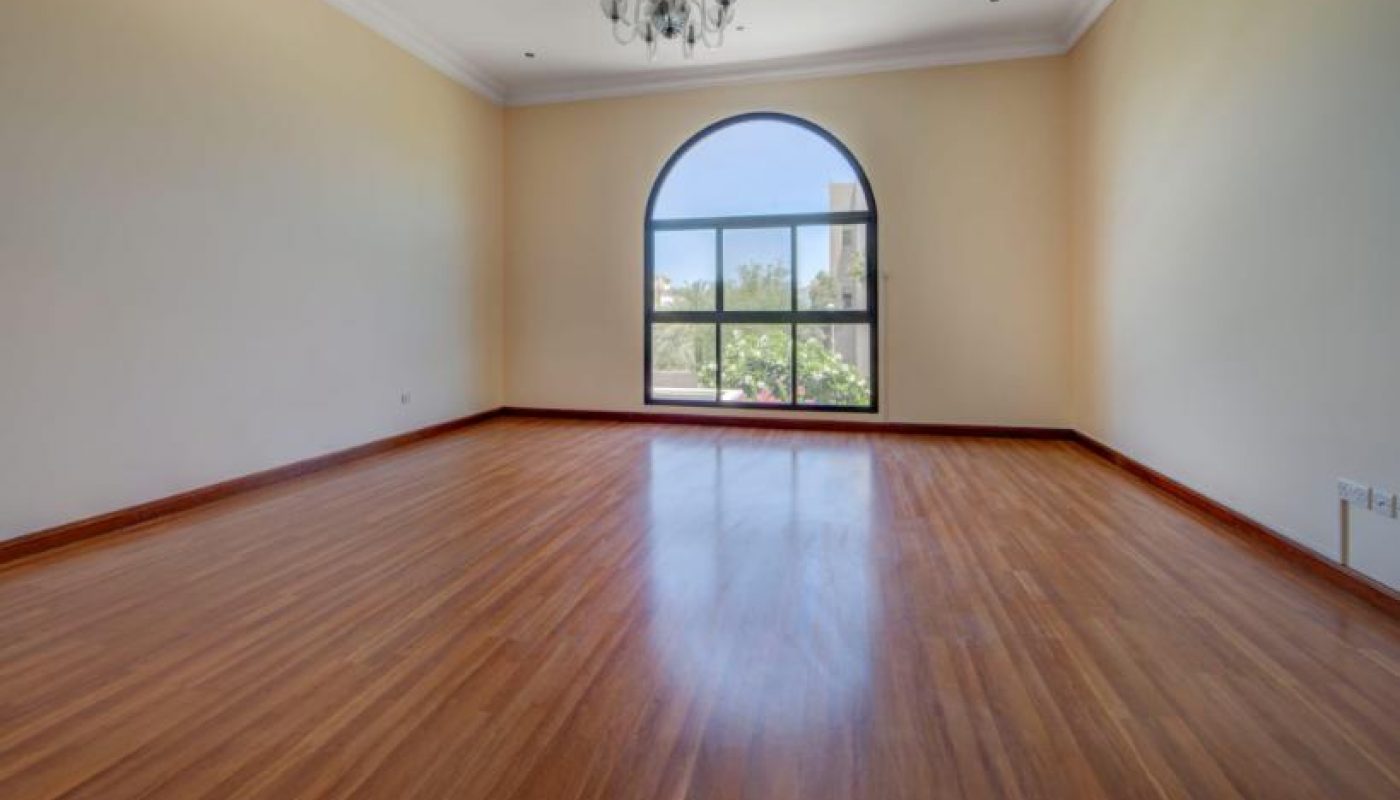 Empty room with polished wooden floor, cream walls, a chandelier, and a large arched window showing an outdoor view.