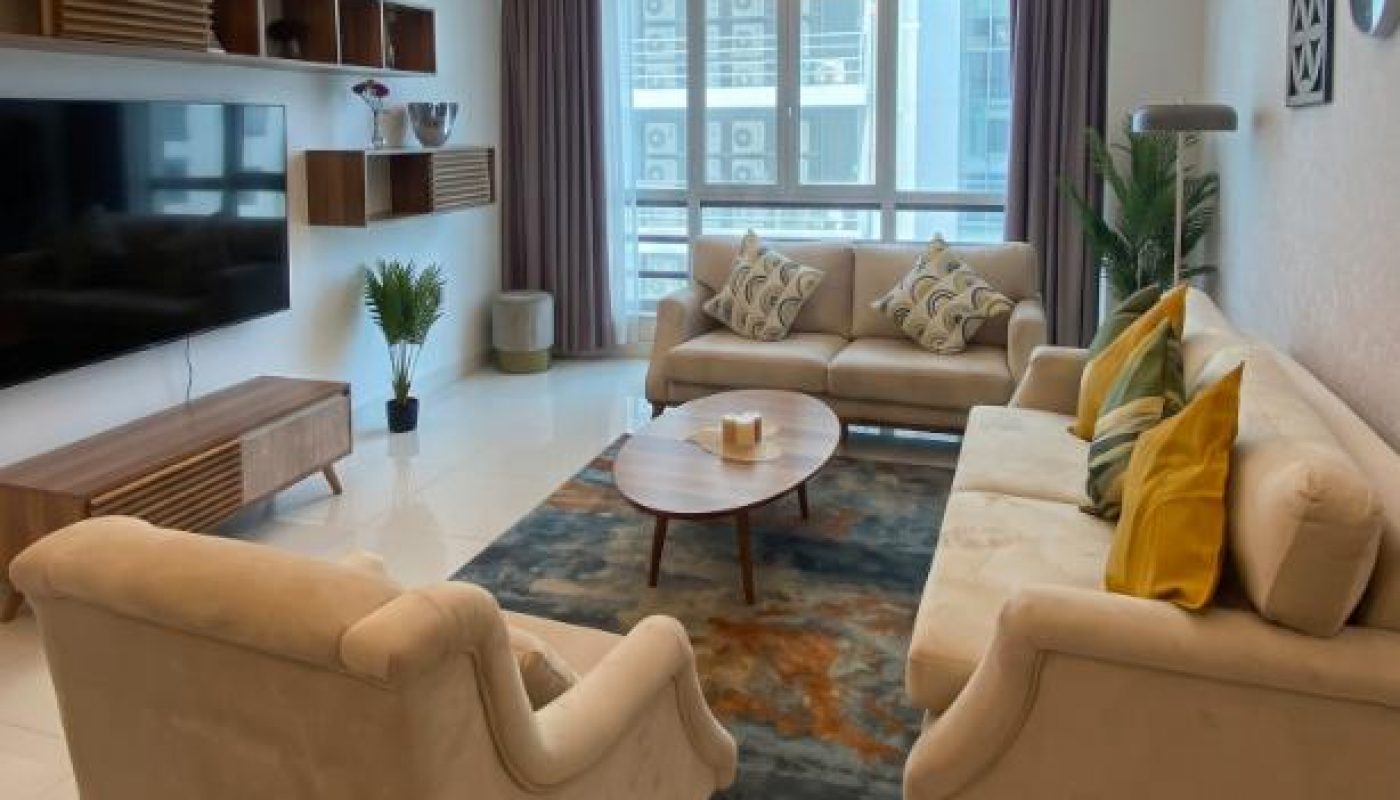 Modern living room with two beige sofas, an armchair, coffee table, wall-mounted TV, decorative shelves, potted plant, and large window with city view.
