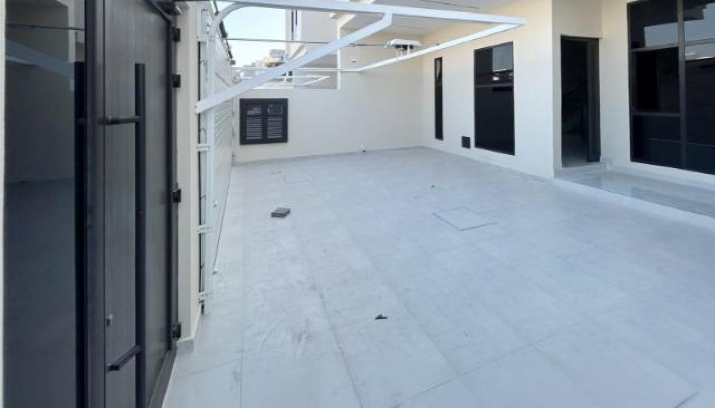 A spacious, modern outdoor patio with white tiles, large glass windows, and sliding doors attached to a contemporary building under clear blue sky.