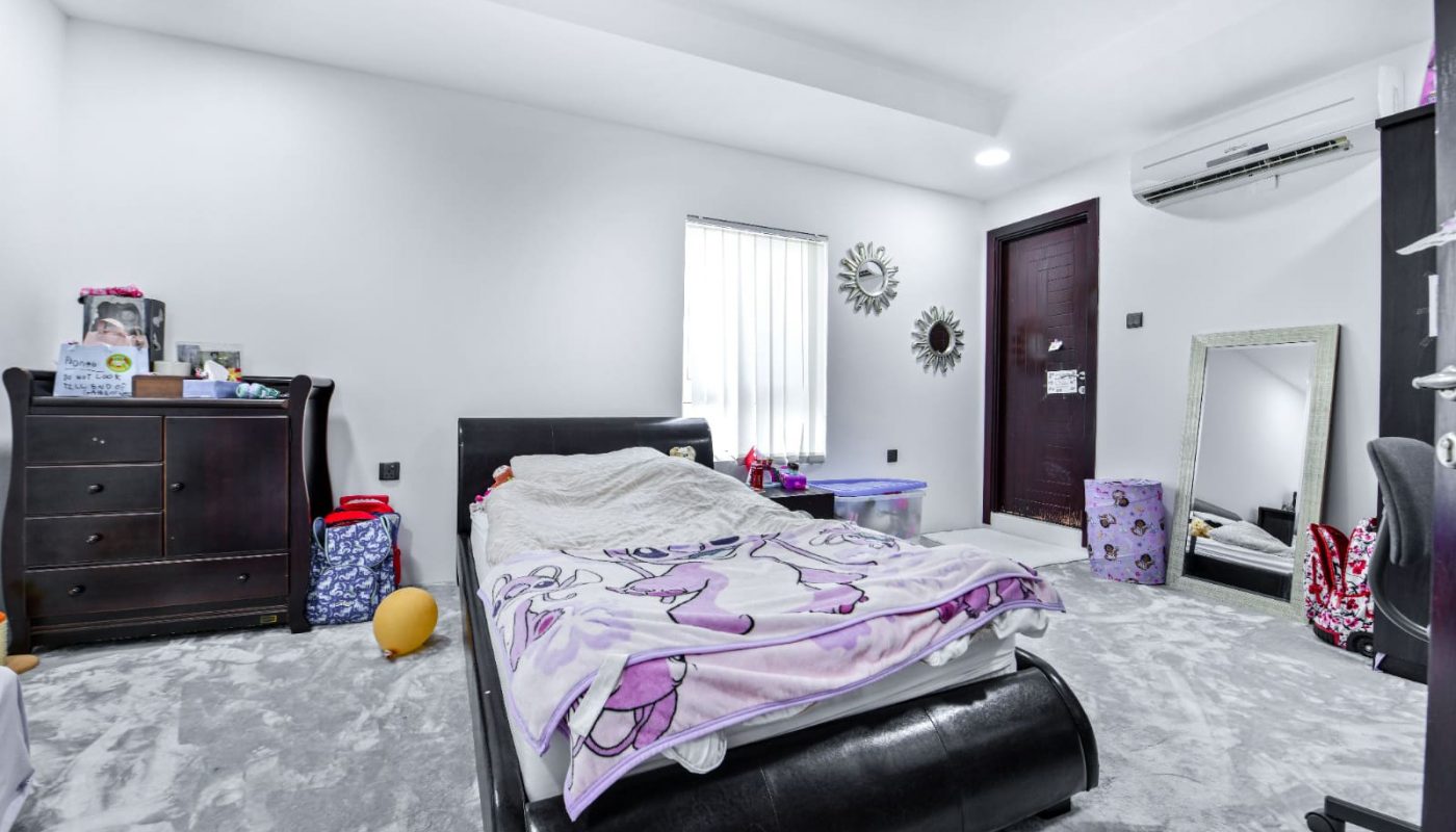 A bedroom with a floral-patterned bed, dark furniture, wall-mounted mirror, air conditioner, and window partially covered by blinds. Items are scattered on the floor and dresser.