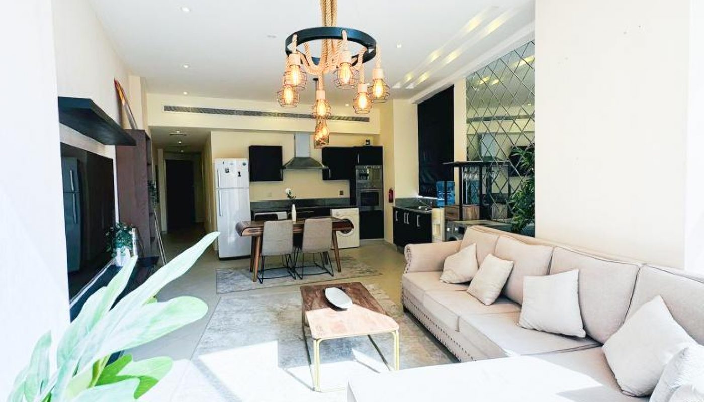 Bright, modern living room with light-colored sectional sofa, dining table, hanging pendant lights, open kitchen, and large window letting in natural light.