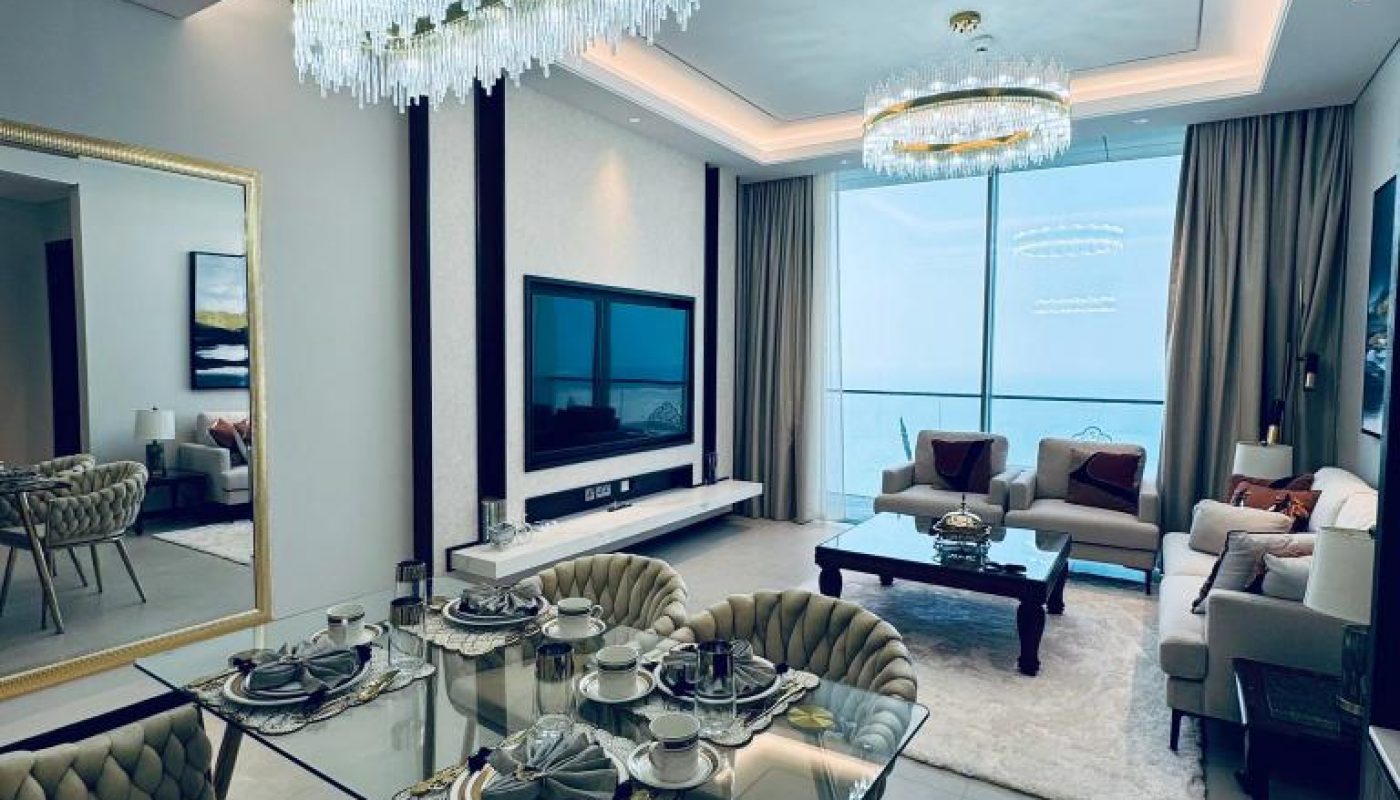 Modern dining and living area with glass table, tufted chairs, large wall mirror, chandelier lighting, and floor-to-ceiling windows with a city view.