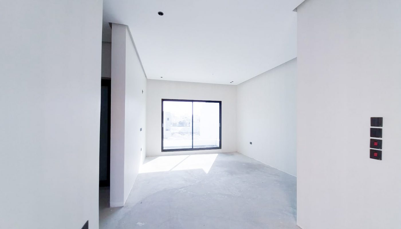 A bright, empty room with white walls, a large floor-to-ceiling window, and an unfinished concrete floor. Four black switches are seen on the right wall.