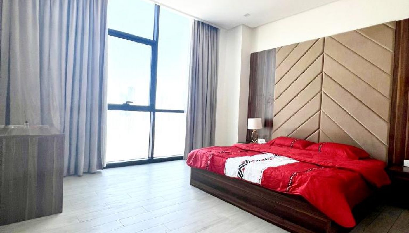 Modern bedroom with a large window, light wood flooring, gray curtains, and a bed with a red comforter next to a nightstand and lamp.