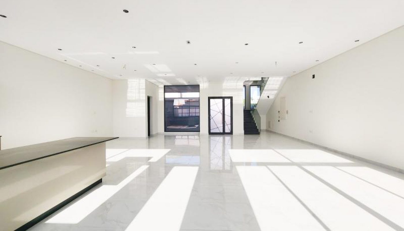 Spacious, empty room with white walls and marble floors, large windows casting shadows, a countertop on the left, and stairs leading up at the back.