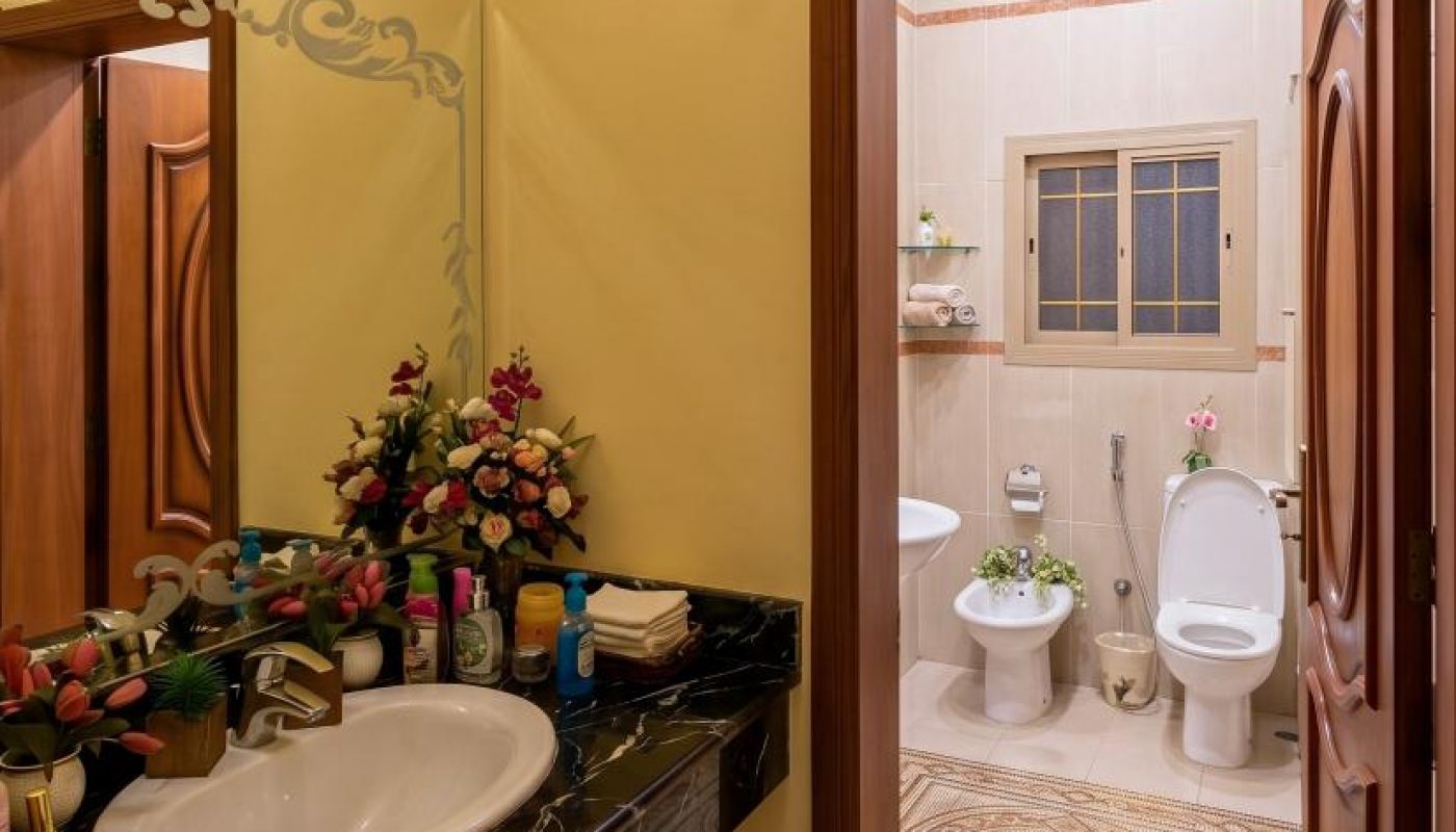A bathroom with a marble countertop sink, a large mirror, toiletries, and flowers; beyond the open door are a toilet, bidet, urinal, and tiled floor.