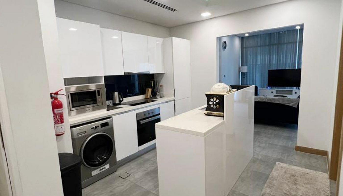 Modern kitchen with white cabinets, an island, built-in appliances, a washing machine, fire extinguisher, and a view into a living area with a TV and a large window.