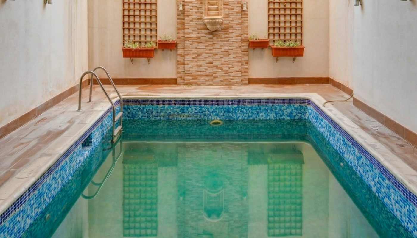 Rectangular indoor swimming pool with tiled blue edges, metal ladder, beige stone walls, and decorative wall features at the far end.