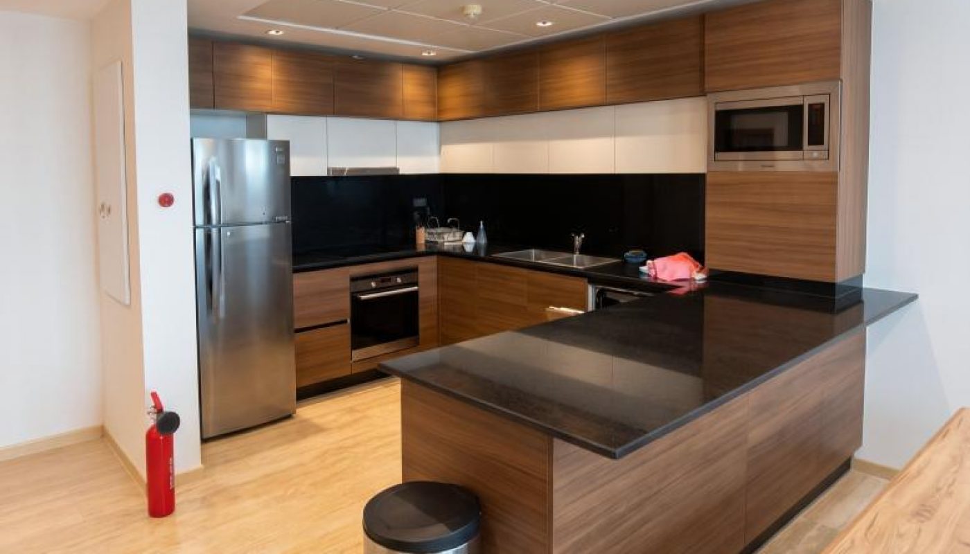 Modern kitchen with wood cabinets, black countertops, stainless steel appliances, a trash can, and a fire extinguisher on a light wood floor. A pink towel is on the counter.