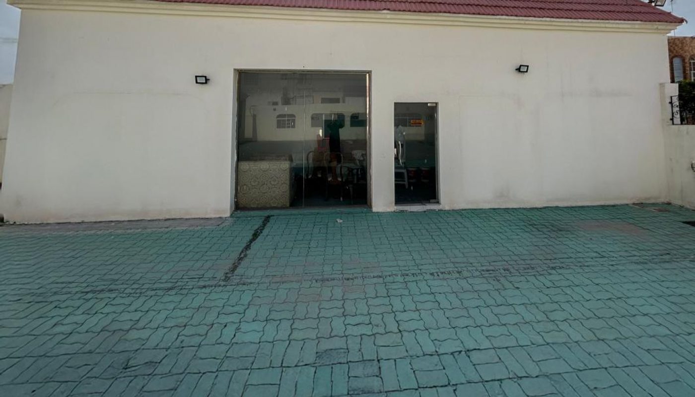 A white building with a red roof and two open doorways is seen under a bright sun, with a paved turquoise courtyard in the foreground.