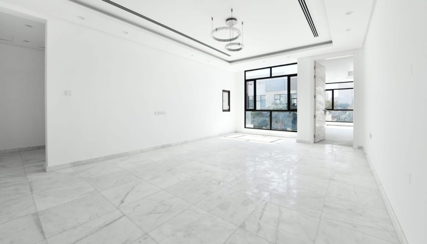 A spacious, empty room with white marble floors, white walls, and a modern light fixture. Large windows on one side provide natural light, and an open doorway leads to another room.