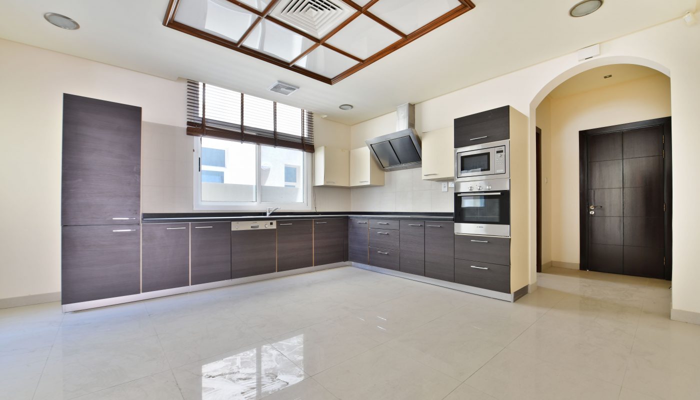 Modern kitchen in a luxury villa with glossy floor tiles, dark wood cabinets, built-in appliances, and a unique wooden ceiling frame, in a bright, empty room.