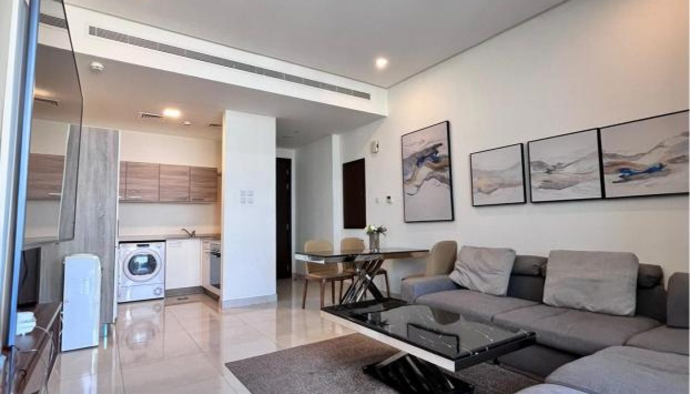 Modern living room with gray sectional sofa, coffee table, wall art, and open kitchen area with light wood cabinets and washing machine. White tile flooring throughout.