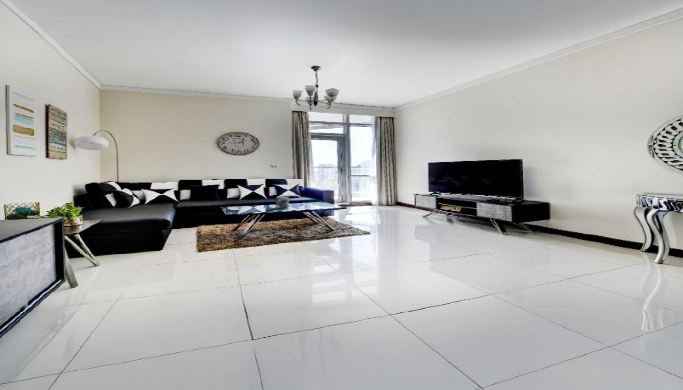 Modern living room with white tiled floor, black sofa, TV, coffee table, wall art, a clock, and large windows with curtains letting in natural light.