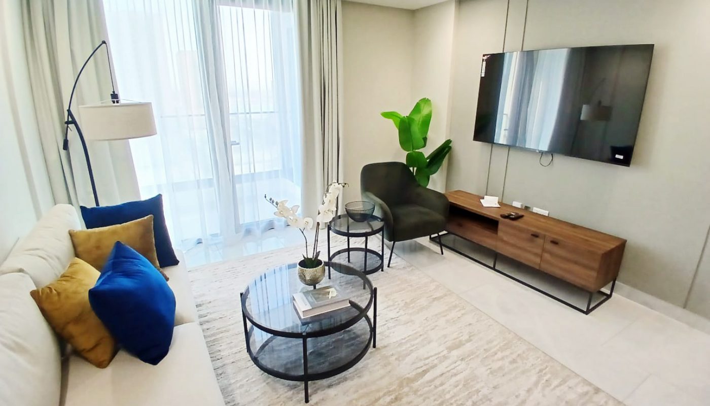 A modern living room in Amwaj Islands features a beige sofa, blue and yellow cushions, glass coffee table, wall-mounted TV, wooden console, and a potted plant—perfect for those seeking stylish freehold apartments for sale.
