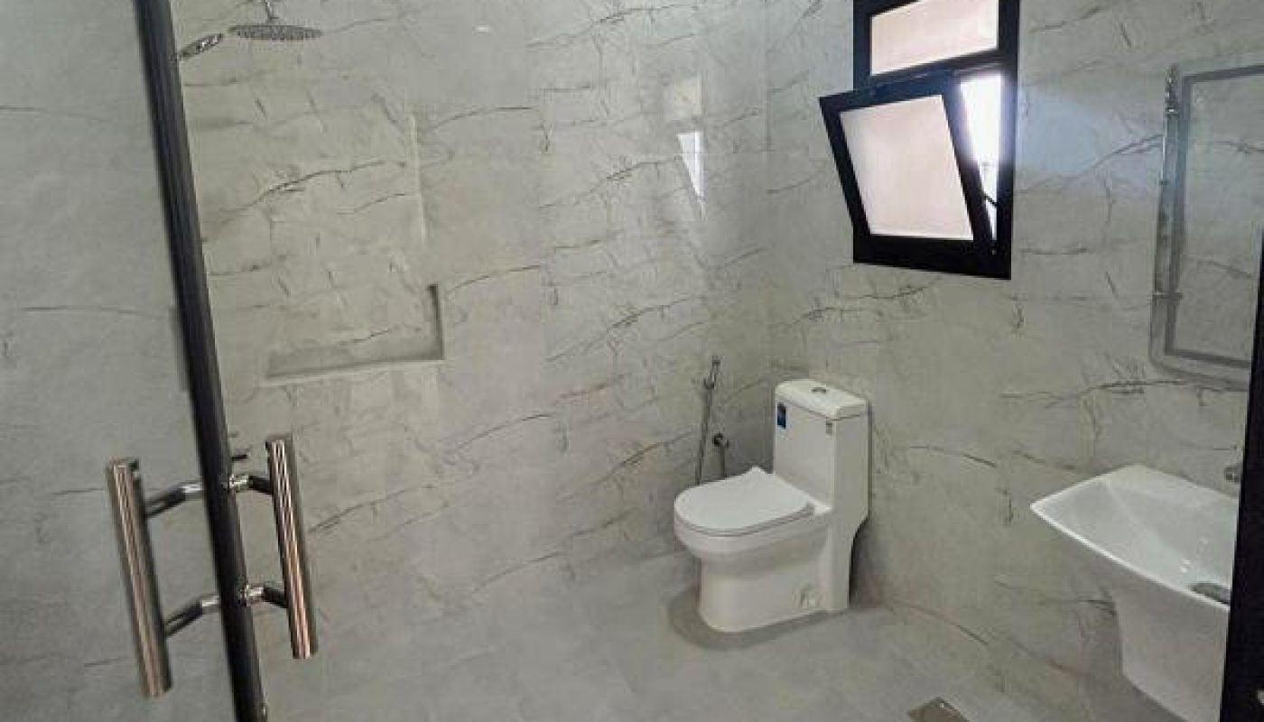 Modern bathroom with gray tiles, a glass-enclosed shower, a white toilet, a small sink, and a window. The room is well-lit with ceiling lights.