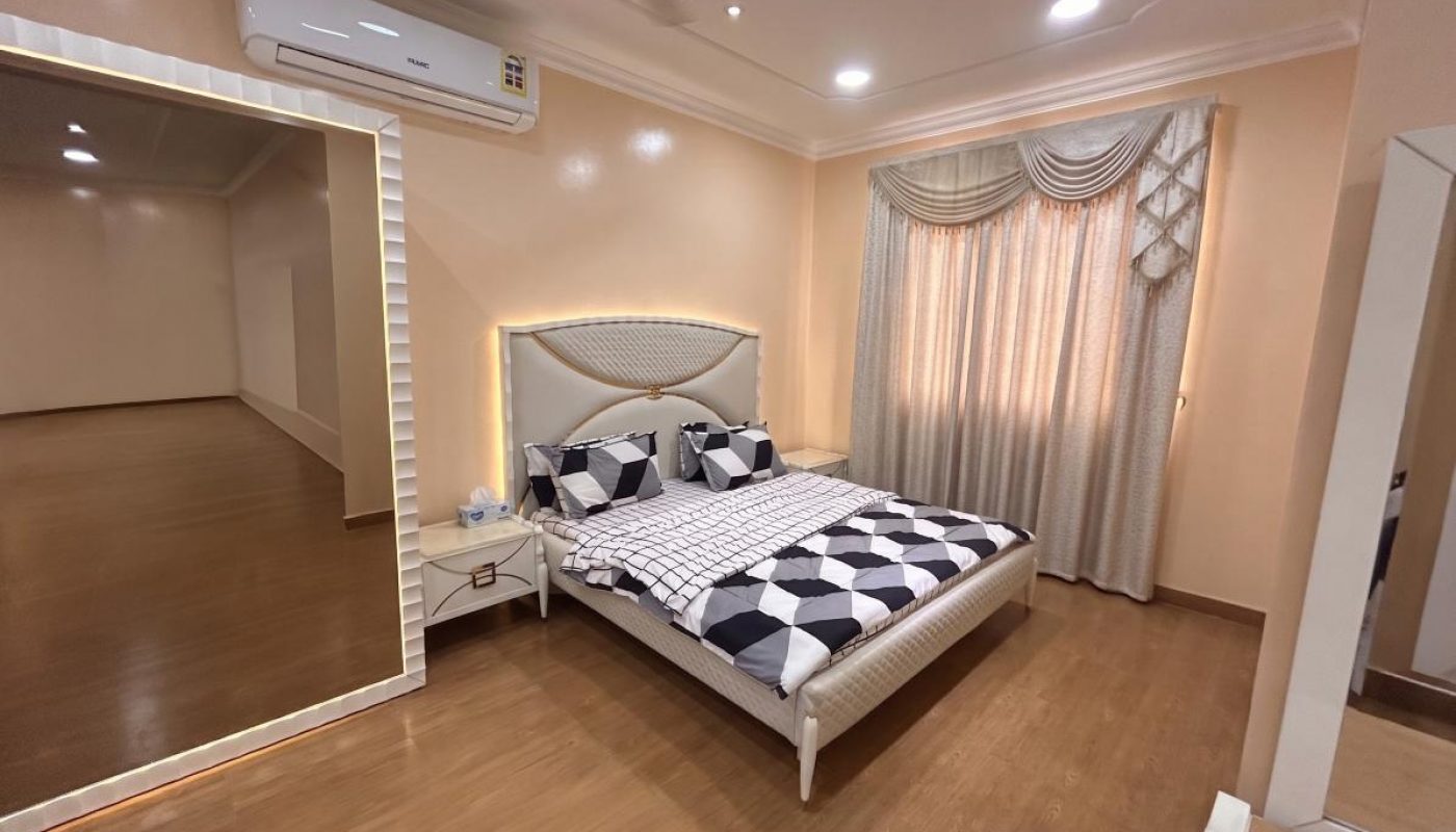 A modern bedroom with a double bed, checkered bedding, nightstand, large wall mirror, beige curtains, wooden floor, and ceiling lights.