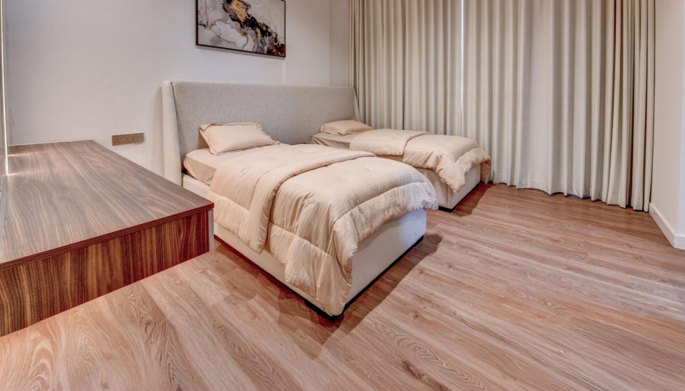 Minimalist bedroom with two single beds, cream bedding, large wooden desk, floor-to-ceiling curtains, abstract wall art, and wood flooring.