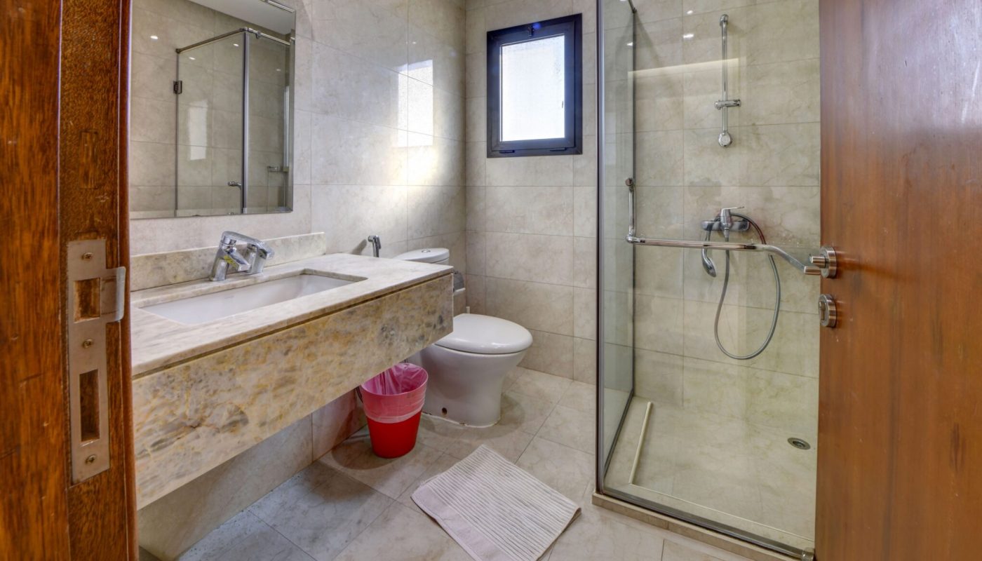 Modern bathroom with a large mirror, dual sink, toilet, red trash can, glass shower enclosure, beige tile walls and floor, and an open wooden door.