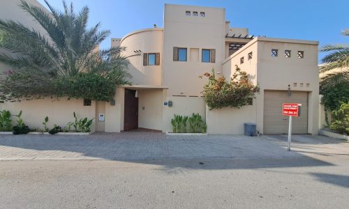 A luxurious villa in Saraya 1, this modern tan two-story house features small windows, a gray garage door, and is surrounded by various plants. With a "For Sale" sign prominently displayed, the driveway and street are also visible.