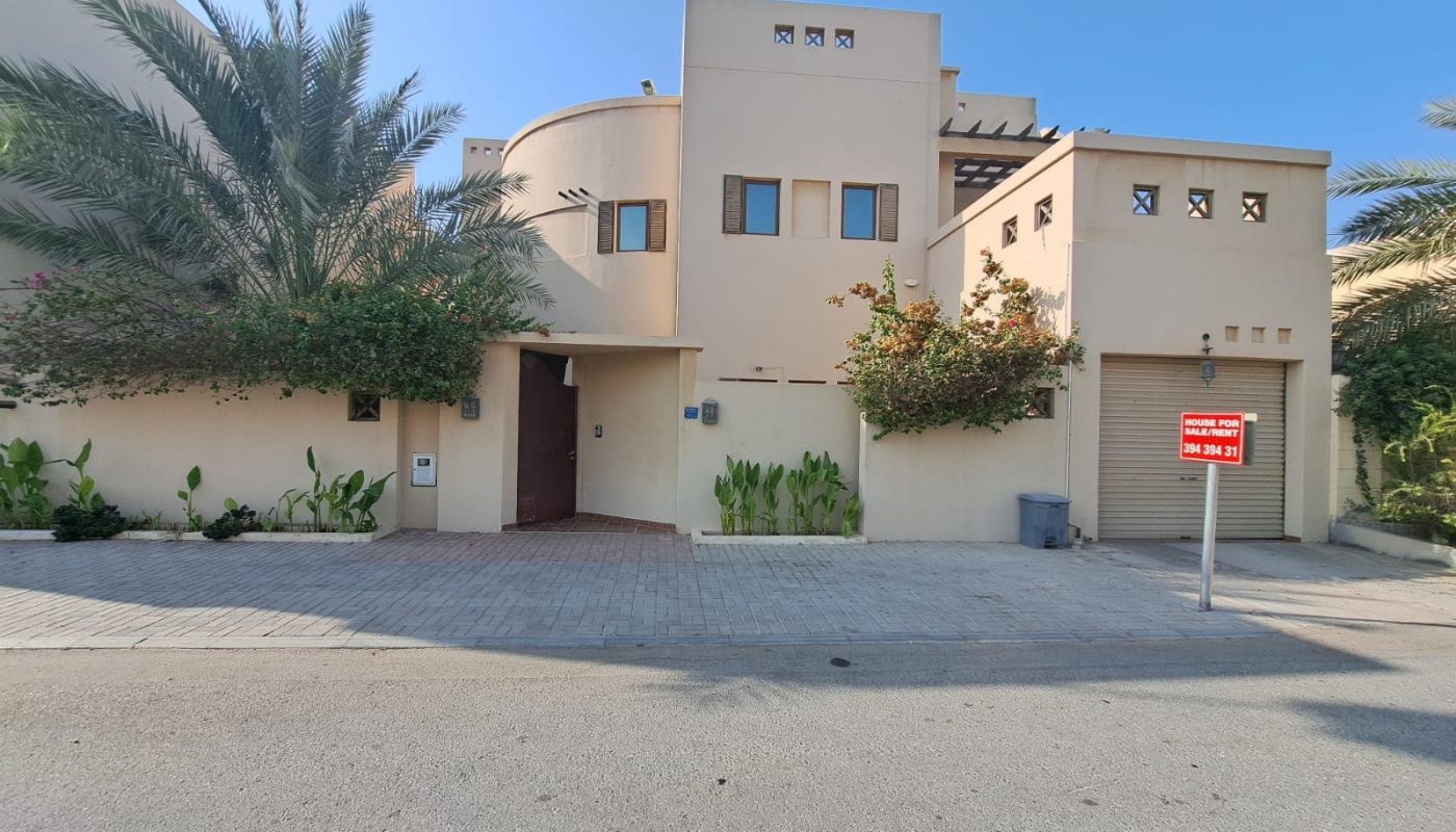 A luxurious villa in Saraya 1, this modern tan two-story house features small windows, a gray garage door, and is surrounded by various plants. With a "For Sale" sign prominently displayed, the driveway and street are also visible.