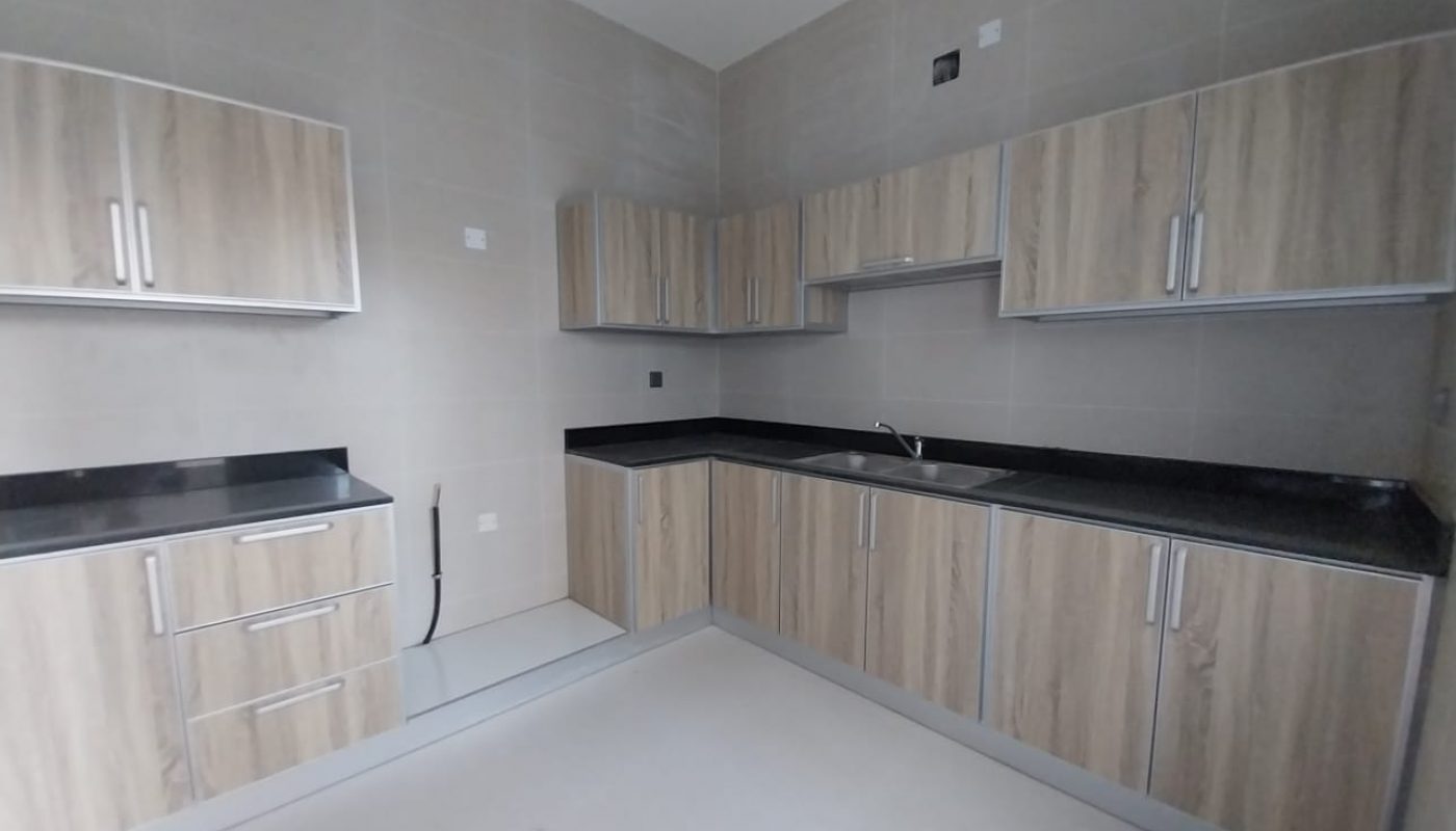 Modern kitchen with light wood cabinets, black countertops, and a stainless steel sink, set against light beige tiled walls and a light floor.