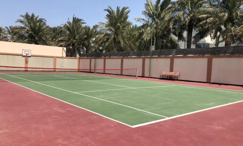 Outdoor tennis court with green playing area surrounded by red surface, set against a backdrop of palm trees and a clear sky.