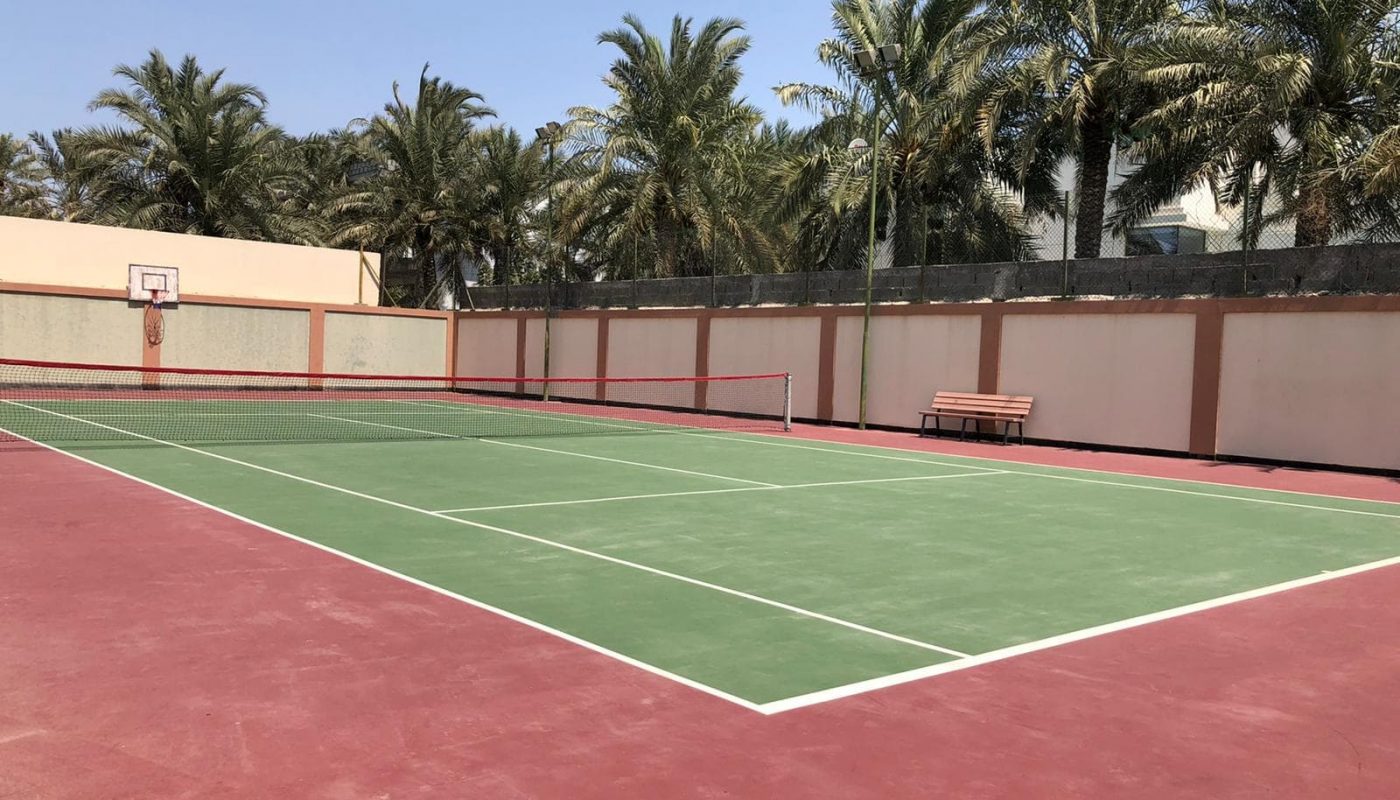 Outdoor tennis court with green playing area surrounded by red surface, set against a backdrop of palm trees and a clear sky.