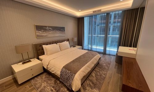 Luxurious apartment with a large bed, patterned rug, artwork above the bed, and natural light from windows with sheer curtains.