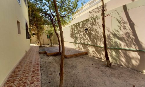 A narrow outdoor area with trees, tiled pathway, and small wooden platforms, located between beige walls of a building under clear skies.