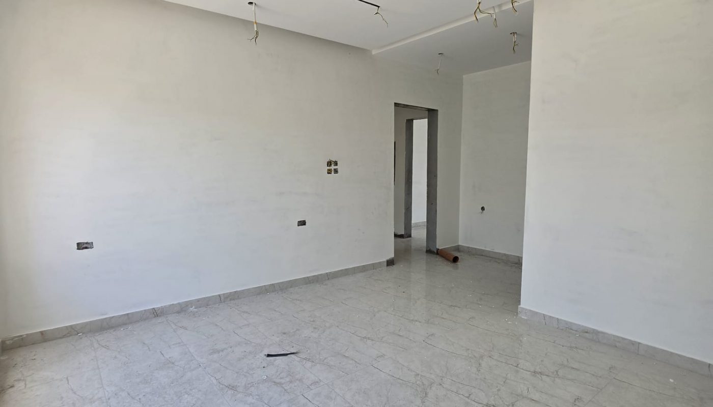 Empty, unfurnished room with light gray tiled floor, plain white walls, exposed ceiling light wires, and an open doorway leading to another area.
