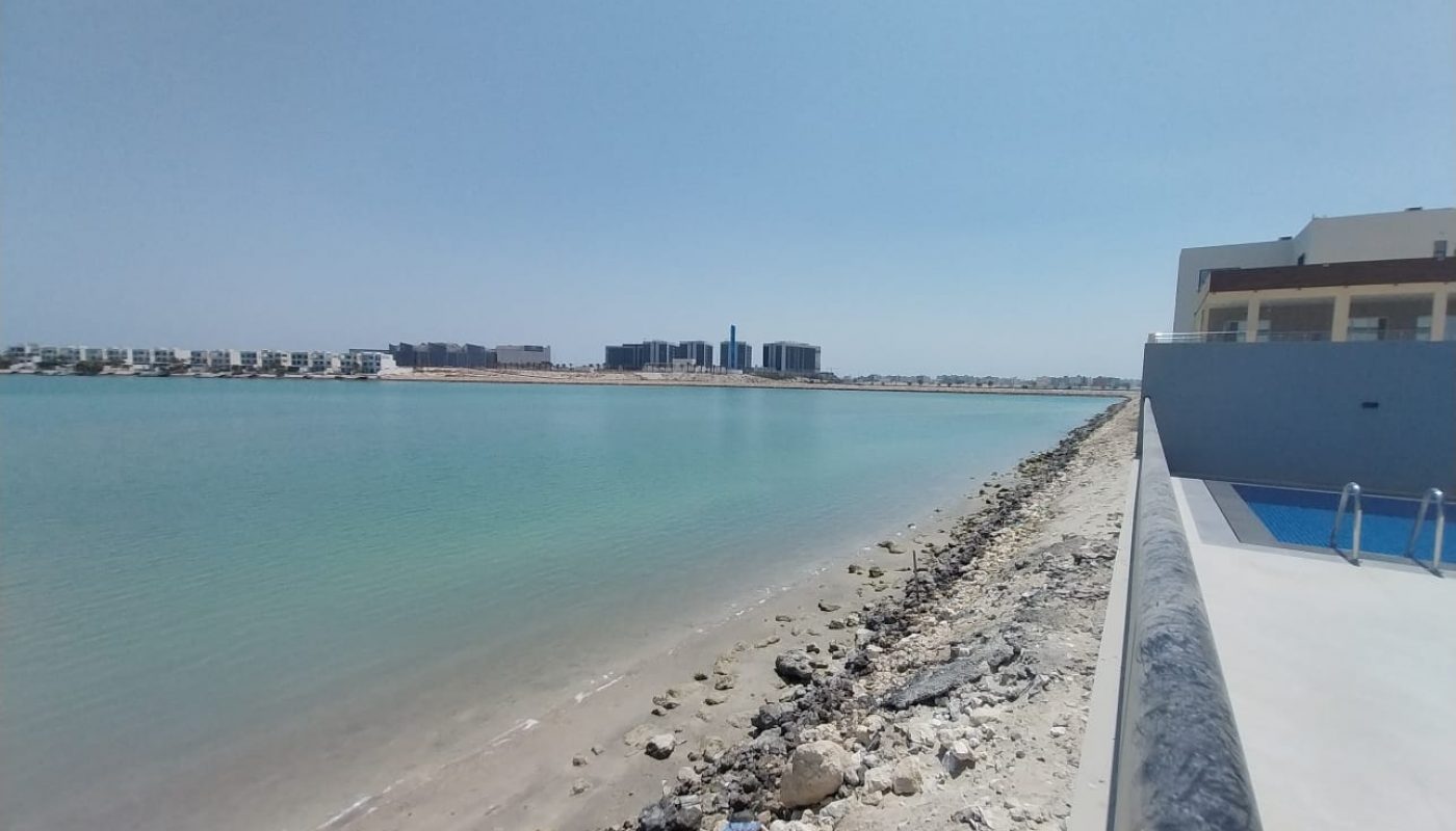 A calm shoreline with clear blue water and pebbles leads to a distant cityscape under a clear sky, with a building on the right.