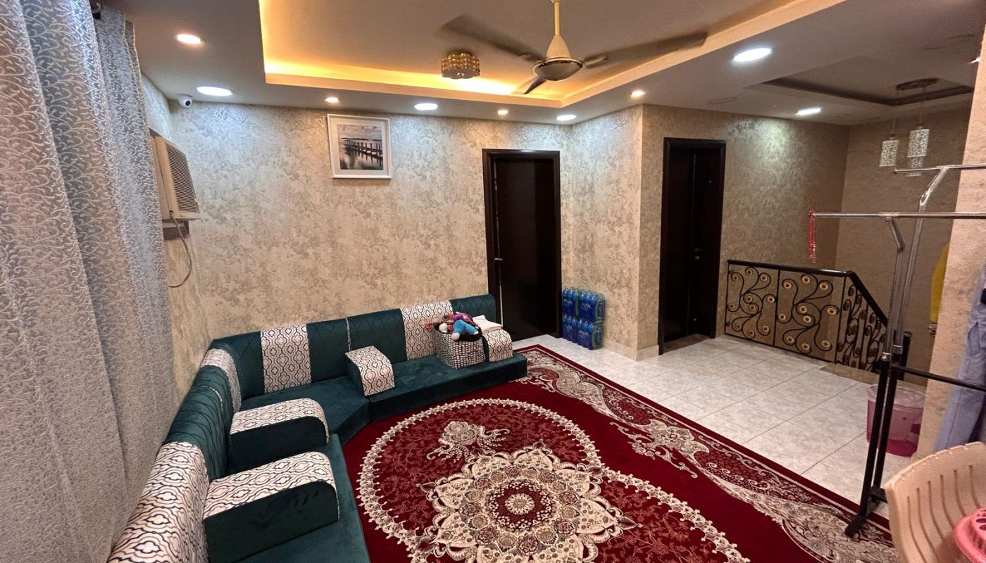 A living room with a patterned red carpet, green and white sectional sofa, ceiling fan, wall art, and staircase with metal railing.