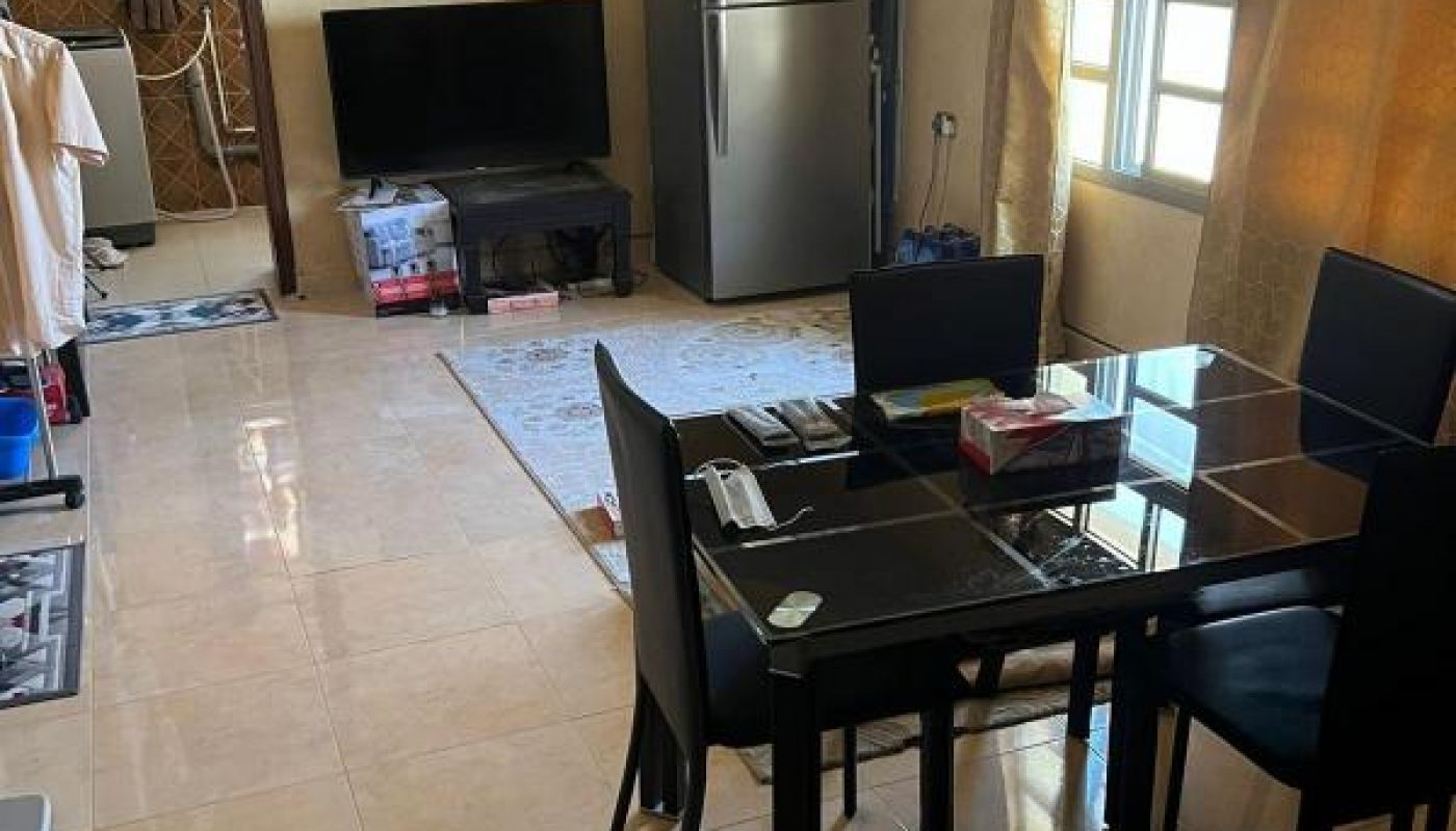 A dining and living area with a glass dining table, black chairs, a TV, refrigerator, and window with golden curtains. Some household items are scattered around the room.