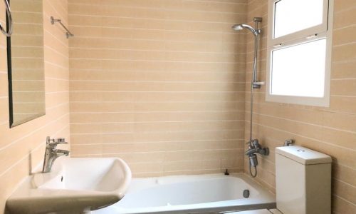 A clean bathroom with beige tiled walls, a white bathtub with a showerhead, a white sink, a toilet, and a window.