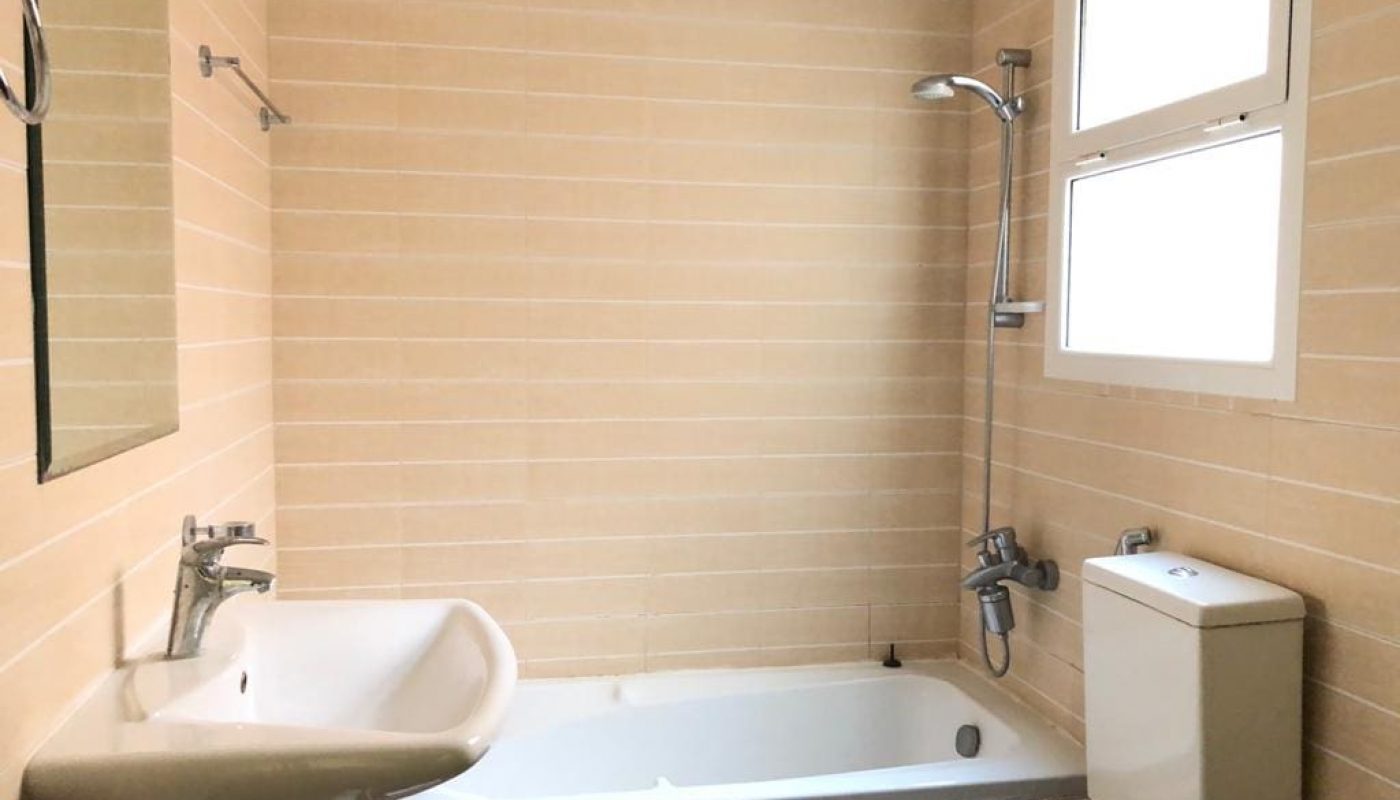 A clean bathroom with beige tiled walls, a white bathtub with a showerhead, a white sink, a toilet, and a window.