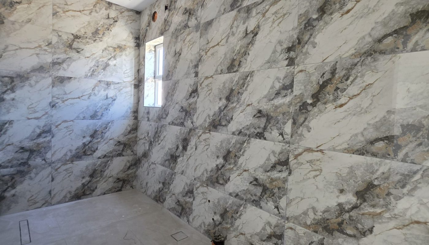 Room under construction with marble-patterned wall tiles, exposed light fixtures, plumbing pipes, and an unfinished floor. A small window provides natural light.