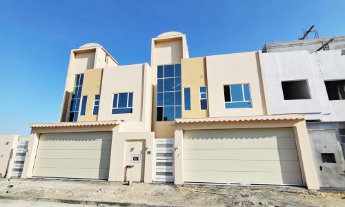 A new beige duplex with large windows and dual auto garages under a clear blue sky.