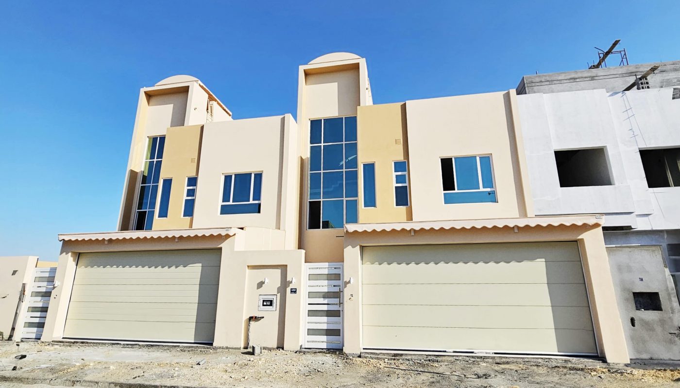 A new beige duplex with large windows and dual auto garages under a clear blue sky.