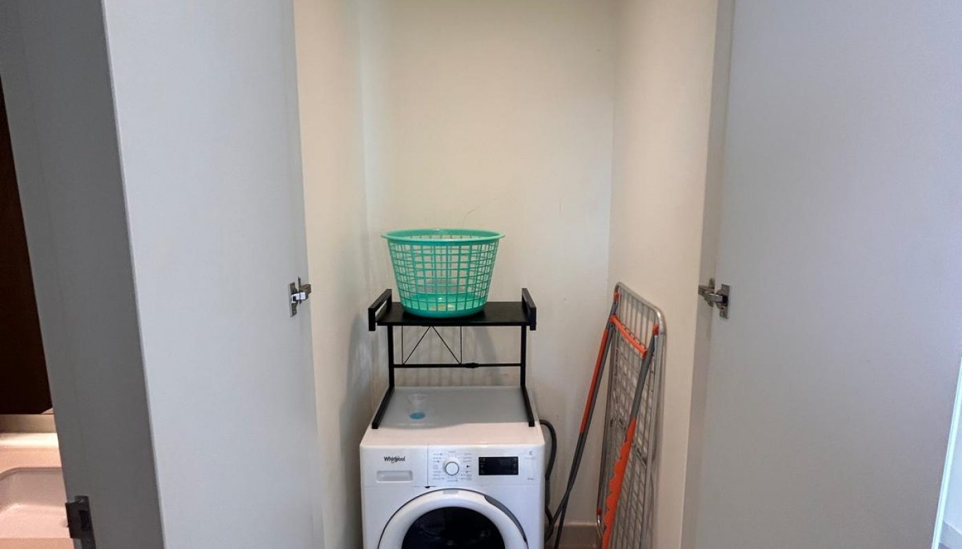 A small laundry closet with a washing machine, green laundry basket, drying rack, a large water bottle, and tiled floor.