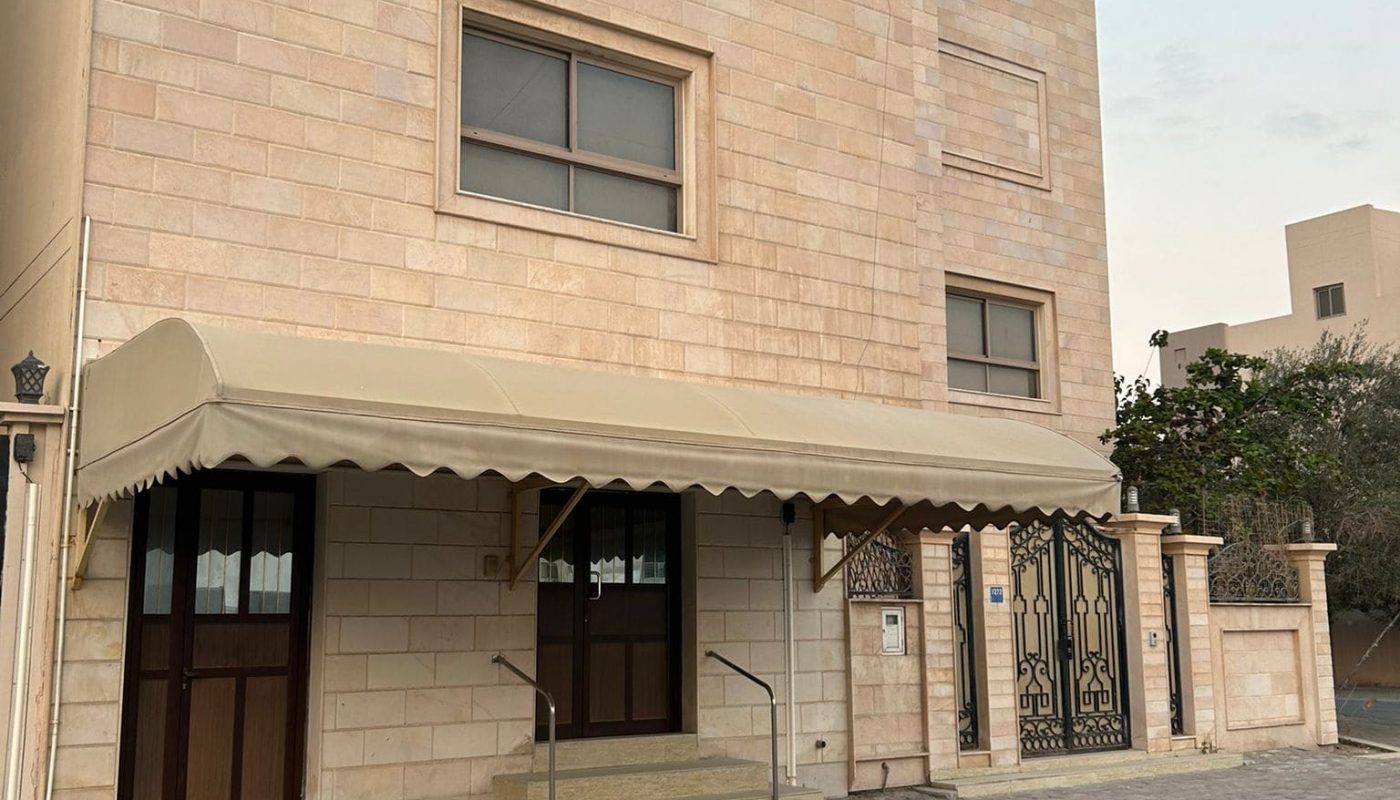 Two-story beige stone building with a large awning over the entrance and a decorative metal gate. The road and sidewalk are in the foreground.