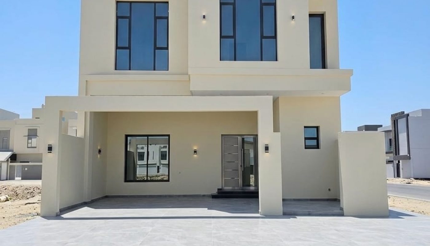Modern two-story beige house with large windows, a flat roof, and a covered driveway, set in a newly developed residential area under a clear sky.