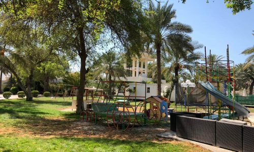 A playground with various equipment including slides, swings, and climbing structures surrounded by grassy areas and trees on a sunny day.