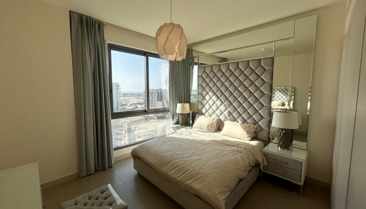 Modern bedroom with a large bed, padded headboard, bedside tables with lamps, a window with city view, and a geometric pendant light.